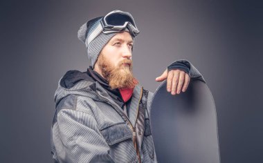 Vahşi kızıl saçlı snowboard uzağa arıyorsunuz bir kış şapka ve koruyucu gözlük bir stüdyoda snowboard ile poz bir snowboard ceket giymiş tam sakallı. Gri arka plan üzerinde izole.