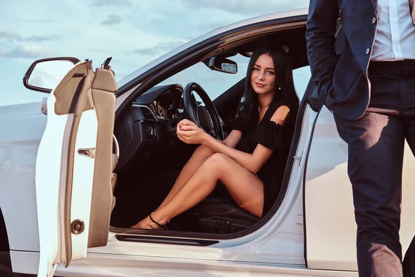 Mujer hermosa bien vestida sentada en el asiento delantero y hombre vestido de traje de pie cerca del coche al aire libre . — Foto de Stock