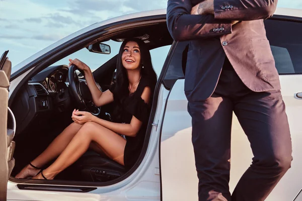 Mujer hermosa bien vestida sentada en el asiento delantero y hombre vestido de traje de pie cerca del coche al aire libre . — Foto de Stock