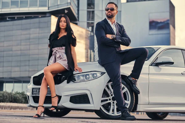 Pareja atractiva bien vestida apoyada en un coche de lujo al aire libre contra el rascacielos . — Foto de Stock