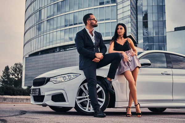 Pareja atractiva bien vestida apoyada en un coche de lujo al aire libre contra el rascacielos . — Foto de Stock