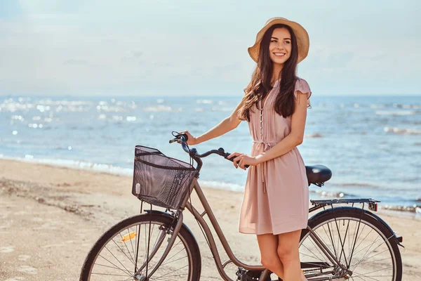 Belle fille brune habillée en robe et chapeau posant avec un vélo sur la plage par une journée ensoleillée . — Photo