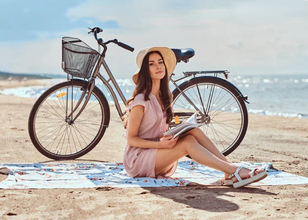 Ung kvinna håller en bok och titta på kameran när du sitter på stranden. — Stockfoto