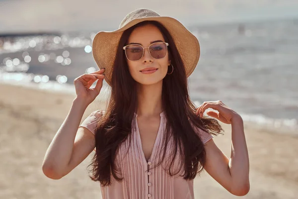 Portret van een lachende mooie brunette meisje met lang haar in zonnebrillen en zonnebril dragen van een jurk op het strand. — Stockfoto