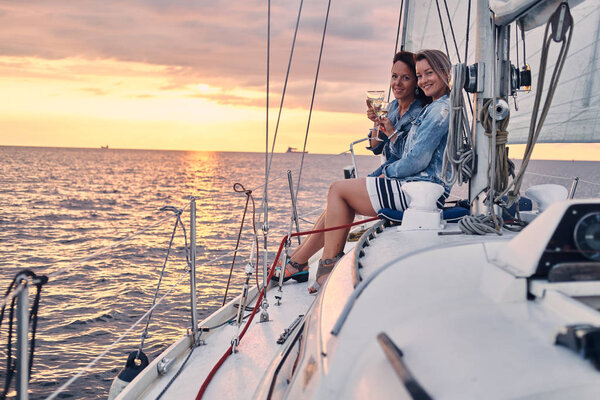 Две подруги отдыхают на яхте с бокалами вина в руках, во время заката в открытом море
.