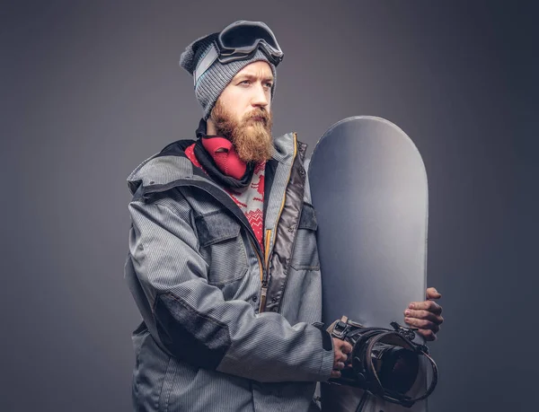 Brutální zrzka snowboardista s plnovousem v zimní čepice a ochranné brýle, oblečený v snowboarding kabát pózuje s snowboard ve studiu, koukal. Na šedém pozadí, samostatný. — Stock fotografie