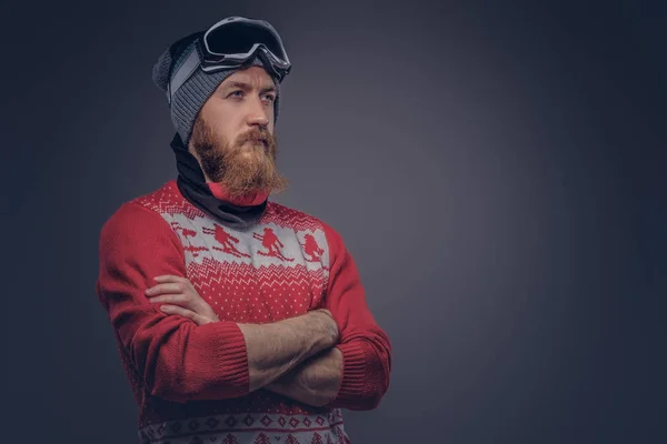 Portrait of a brutal redhead bearded male in a winter hat with protective glasses dressed in a red sweater, posing with crossed arms at a studio. Isolated on the gray background. — Stock Photo, Image