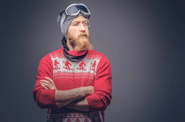 Porträt eines brutalen rothaarigen, bärtigen Mannes mit Wintermütze, Schutzbrille und rotem Pullover, der mit verschränkten Armen in einem Studio posiert. isoliert auf dem grauen Hintergrund. — Stockfoto