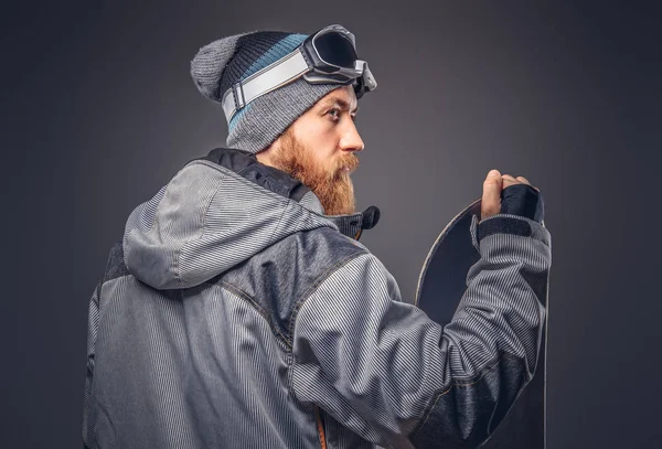 Brutal redhead snowboarder with a full beard in a winter hat and protective glasses dressed in a snowboarding coat posing with snowboard at a studio, looking away. Isolated on the gray background. — Stock Photo, Image