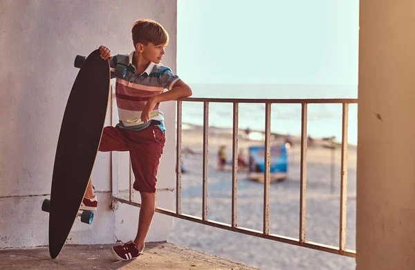 Portrét mladý skejťák na sobě tričko a šortky, opírali se o zábradlí a drží skateboard na pozadí pobřeží na západ slunce. — Stock fotografie