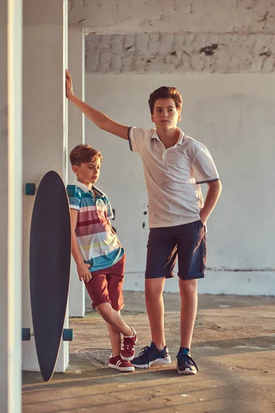 Retrato de dos hermanos pequeños con un monopatín apoyado en una pared al aire libre al atardecer . —  Fotos de Stock