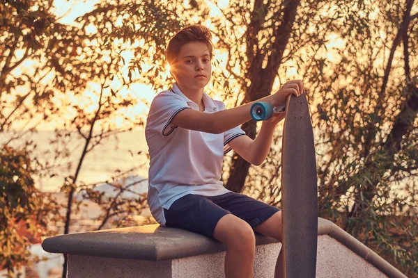 Mladý skejťák na sobě tričko a šortky sedí na kamenné zábradlí venku na západ slunce. — Stock fotografie