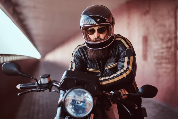 Retrato de cerca de un brutal motorista barbudo con casco y gafas de sol vestido con una chaqueta de cuero negro sentado en una motocicleta retro con un faro incluido . — Foto de Stock