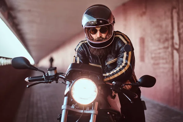 Close-up portrait of a brutal bearded biker in helmet and sunglasses dressed in a black leather jacket sitting on a retro motorcycle with an included headlight. — Stock Photo, Image