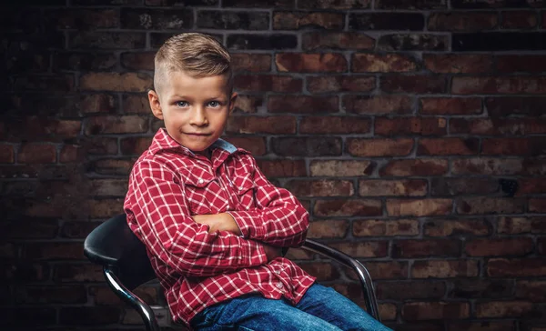 Portrét stylový malého chlapce oblečená v košili a džínách, sedí v křesle proti cihlové zdi. — Stock fotografie