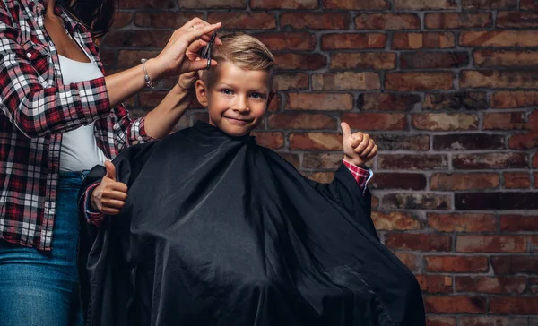 Mutlu sevimli okul öncesi çocuk başparmak bir saç kesimi elde ederken ortaya çıktı. Çocuk Kuaför makası ve tarak ile çatı iç odadaki küçük çocuk kesiyor. — Stok fotoğraf