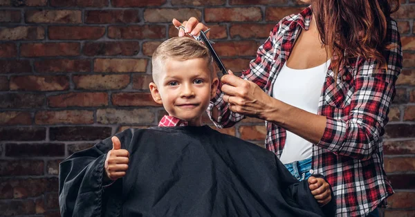 Contented cute preschooler boy shows thumbs up while getting a haircut. Children hairdresser with scissors and comb is cutting little boy in the room with loft interior. — Stock Photo, Image