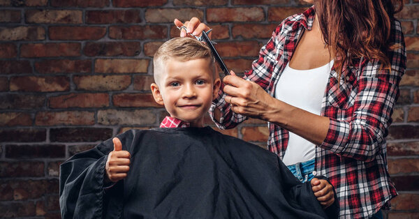 Contented cute preschooler boy shows thumbs up while getting haircut. Children hairdresser with scissors and comb is cutting little boy in the room with loft interior.