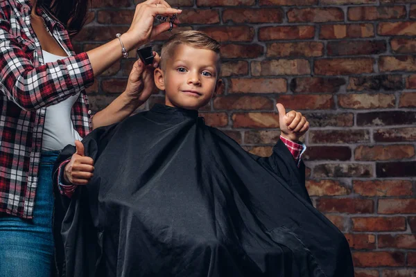 Nöjd söt förskolebarn pojke visar tummen samtidigt få en frisyr. Barn Frisör med sax och kam skär liten pojke i rummet med loft interiör. — Stockfoto