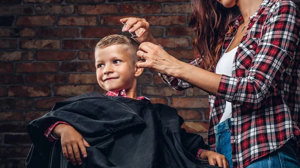 Улыбающийся дошкольник стрижется. Детский парикмахер с ножницами и расческой режет маленького мальчика в комнате с интерьером лофта . — стоковое фото