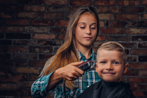 Smiling preschooler boy getting haircut. The older sister cuts her little brother with a trimmer against a brick wall.