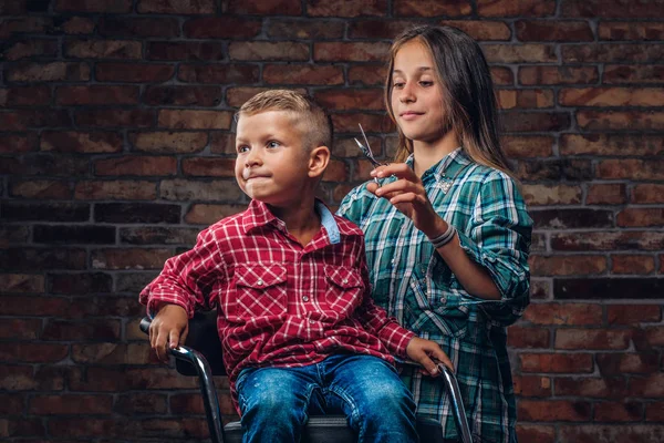 Roztomilý preschooler chlapec u holiče. Starší sestra kusy svého bratříčka s nůžkami proti cihlové zdi. — Stock fotografie