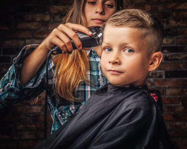 Close-up portrait of a cute preschooler boy getting haircut. The older sister cuts her little brother with a trimmer against the brick wall. — Stock Photo, Image