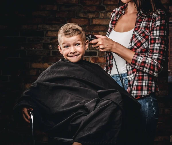 Смішний хлопчик дошкільнята отримує зачіску. Дитячий перукар з тримером ріже маленького хлопчика в кімнаті з інтер'єром лофт . — стокове фото