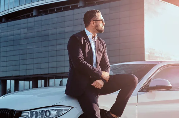 Красивый бородатый мужчина в солнцезащитных очках, одетый в черный костюм, сидит на роскошной машине напротив небоскреба . — стоковое фото