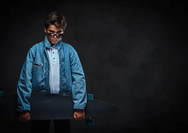 Módní mladý muž v sluneční brýle oblečen v tričku a džínové sako drží longboard. — Stock fotografie