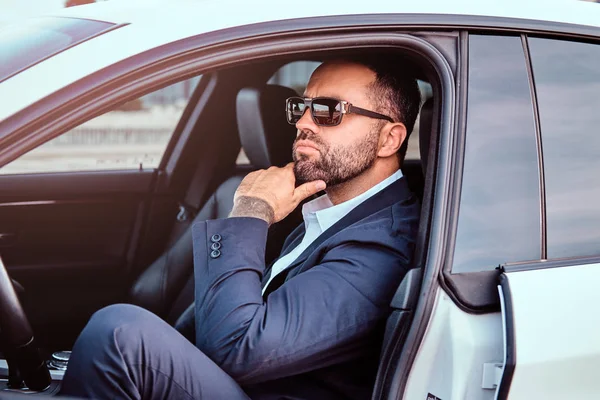 Empresário confiante em óculos de sol vestido em um terno formal sentado nos assentos da frente no carro de luxo . — Fotografia de Stock