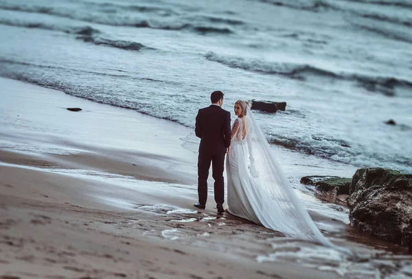 Casamento casal segura uns aos outros mãos andando ao longo da praia perto de ondas do oceano na luz da noite . — Fotografia de Stock
