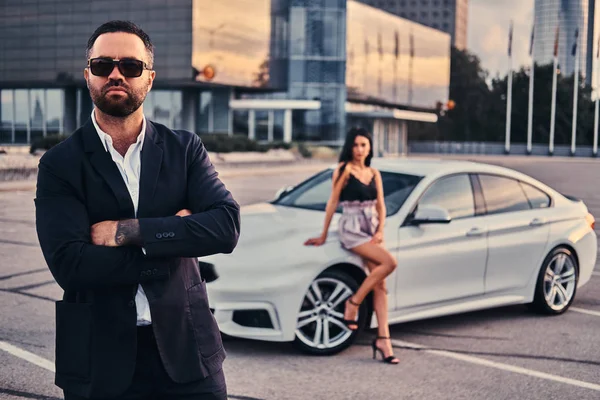 Pareja atractiva bien vestida apoyada en un coche de lujo al aire libre contra el rascacielos . — Foto de Stock