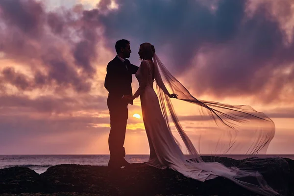 Bröllop par håller varandra händerna stående på stranden mot den fantastiska solnedgången. — Stockfoto