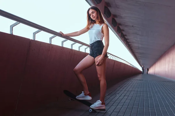 Young sensual girl dressed in shorts and t-shirt standing on a skateboard while leaning on the guardrail at sidewalk under the bridge. — Stock Photo, Image