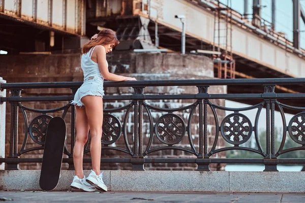 Back view of a sensual skater girl dressed in shorts and t-shirt leaning on the guardrail at a embankment. — Stock Photo, Image
