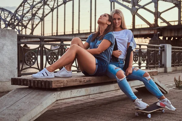 Two beautiful hipster girls sitting on the bench on a background of the skyscraper. — Stock Photo, Image