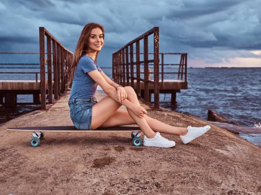 Sahilde bir kaykay üzerinde oturan şehvetli kız inanılmaz karanlık bulutlu hava günbatımı sırasında keyif alıyor.