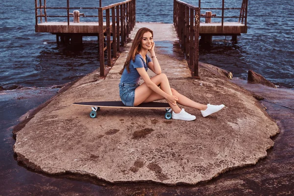 Retrato de uma menina sentada em um skate no cais velho está desfrutando de incrível clima nublado escuro durante o pôr do sol . — Fotografia de Stock