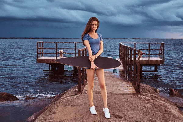 Porträtt av en sensuell tjej håller en skateboard medan stående på stranden njuter av fantastiska mörka molnigt väder under solnedgången. — Stockfoto
