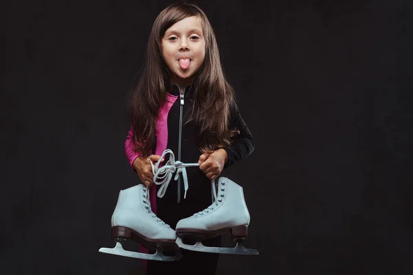 Das verspielte kleine Mädchen in Sportkleidung hält Schlittschuhe in der Hand und zeigt die herausgerissene Zunge. isoliert auf dunkel strukturiertem Hintergrund. — Stockfoto