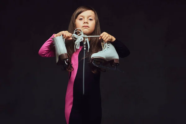Serious little girl dressed in sportswear holds ice skates. Isolated on a dark textured background. — Stock Photo, Image