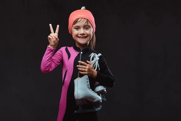 Fröhliches kleines Eisläufermädchen in Sportbekleidung hält Schlittschuhe auf einer Schulter und zeigt Siegerhand. isoliert auf dunkel strukturiertem Hintergrund. — Stockfoto