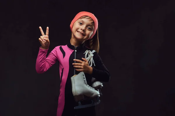 Feliz niña patinadora vestida con ropa deportiva sostiene patines de hielo en un hombro y muestra la mano de la victoria. Aislado sobre fondo de textura oscura . —  Fotos de Stock