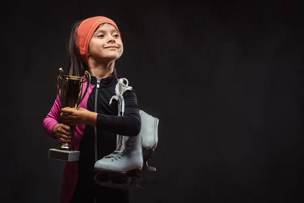 Happy little skater girl champion holds a winners cup and ice skates. Isolated on dark textured background.