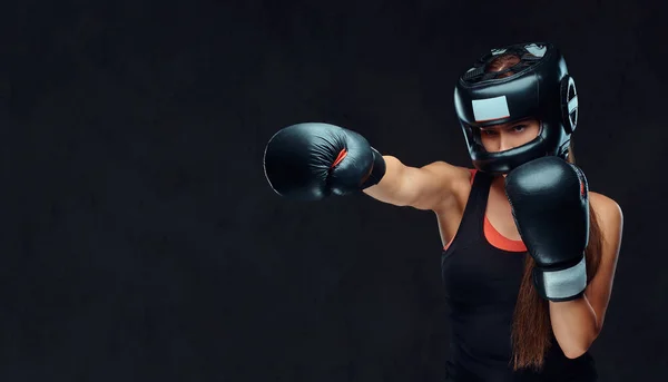 Αθλητική γυναίκα σε αθλητικά ενδύματα που φορώντας ένα προστατευτικό κράνος και γάντια του μποξ, κατάρτισης στο γυμναστήριο. Απομονωμένη σε ένα σκοτεινό φόντο με υφή. — Φωτογραφία Αρχείου