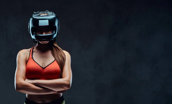 Mulher esportiva em sutiã esportivo usando um capacete protetor, posando com os braços cruzados e olhando para uma câmera. Isolado em fundo escuro texturizado . — Fotografia de Stock