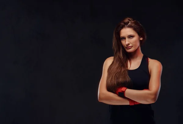 Portrait of a beautiful sportive female boxer in sportswear and bandaged hands, posing with crossed arms. Isolated on dark textured background.