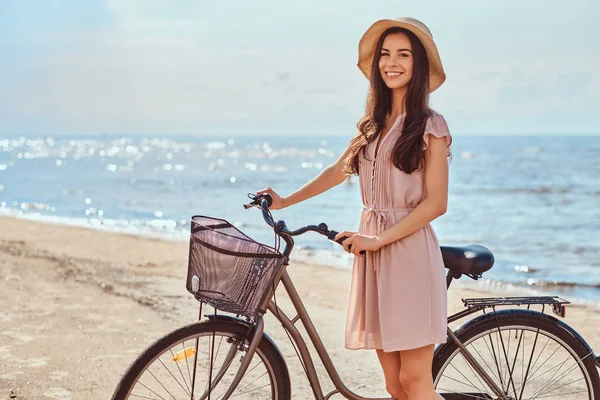 Belle fille brune habillée en robe et chapeau posant avec un vélo sur la plage par une journée ensoleillée . — Photo