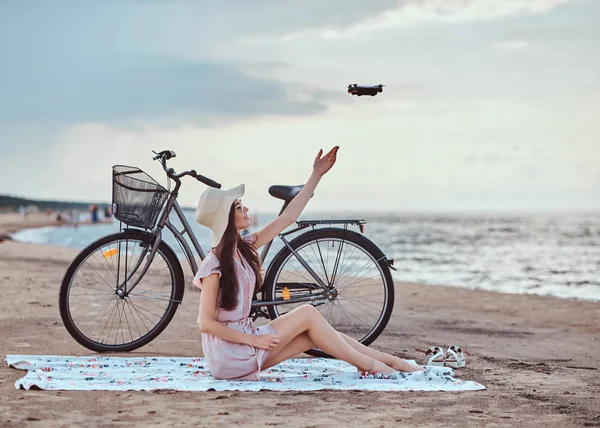 Porträtt av en ung brunett flicka i solglasögon och hatt bär klänning njuta av semestern på stranden, spela med en quadcopter. — Stockfoto